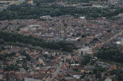 Aerial view of a city with dense historical architecture, featuring church steeples and a mix of modern buildings surrounded by lush green areas. Red and brown rooftops dominate the landscape, and wide streets intersect through the cityscape.