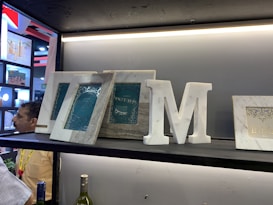 A shelf displays various items including picture frames, a large decorative letter 'M', and a couple of bottles. The frames have an intricate design with 'INDIA' inscribed on them. The setup appears to be part of a display at an exhibition or a store, with a partial view of people in the background.