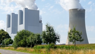 A large industrial facility with multiple tall structures emitting white smoke is situated next to a cooling tower. The facility is surrounded by greenery, including trees and grass, and there is a road running along the side.