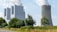 A large industrial facility with multiple tall structures emitting white smoke is situated next to a cooling tower. The facility is surrounded by greenery, including trees and grass, and there is a road running along the side.