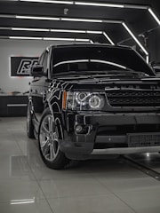 A shiny black Range Rover is parked inside a modern showroom with sleek, reflective surfaces. The vehicle is positioned at an angle with emphasis on its front end, showcasing the headlights and grille. Overhead lighting creates a sophisticated ambiance, highlighting the car's polished exterior.