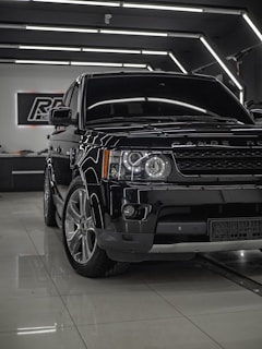 A shiny black Range Rover is parked inside a modern showroom with sleek, reflective surfaces. The vehicle is positioned at an angle with emphasis on its front end, showcasing the headlights and grille. Overhead lighting creates a sophisticated ambiance, highlighting the car's polished exterior.