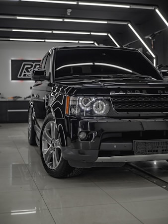 A shiny black Range Rover is parked inside a modern showroom with sleek, reflective surfaces. The vehicle is positioned at an angle with emphasis on its front end, showcasing the headlights and grille. Overhead lighting creates a sophisticated ambiance, highlighting the car's polished exterior.