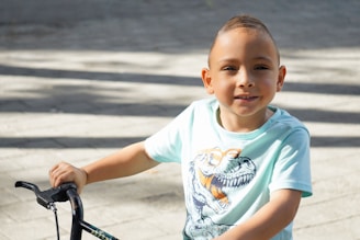 A young child is sitting on a bicycle, wearing a light blue shirt featuring a dinosaur print with glasses. The child has short hair and is smiling gently, with natural sunlight casting shadows on a paved surface.