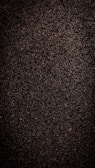 A close-up view of dark, coarse textured asphalt with a rough and uneven surface. The material appears dense with small stones and pebbles visible throughout.