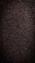 A close-up view of dark, coarse textured asphalt with a rough and uneven surface. The material appears dense with small stones and pebbles visible throughout.