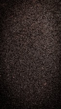 A close-up view of dark, coarse textured asphalt with a rough and uneven surface. The material appears dense with small stones and pebbles visible throughout.