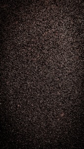 A close-up view of dark, coarse textured asphalt with a rough and uneven surface. The material appears dense with small stones and pebbles visible throughout.