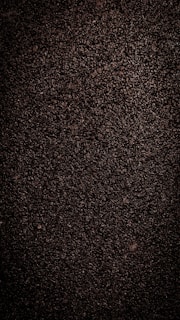 A close-up view of dark, coarse textured asphalt with a rough and uneven surface. The material appears dense with small stones and pebbles visible throughout.