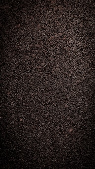 A close-up view of dark, coarse textured asphalt with a rough and uneven surface. The material appears dense with small stones and pebbles visible throughout.