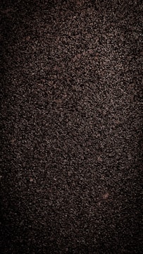 A close-up view of dark, coarse textured asphalt with a rough and uneven surface. The material appears dense with small stones and pebbles visible throughout.