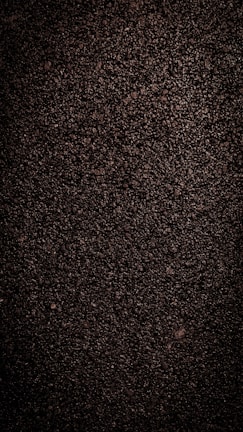A close-up view of dark, coarse textured asphalt with a rough and uneven surface. The material appears dense with small stones and pebbles visible throughout.