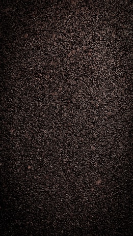 A close-up view of dark, coarse textured asphalt with a rough and uneven surface. The material appears dense with small stones and pebbles visible throughout.