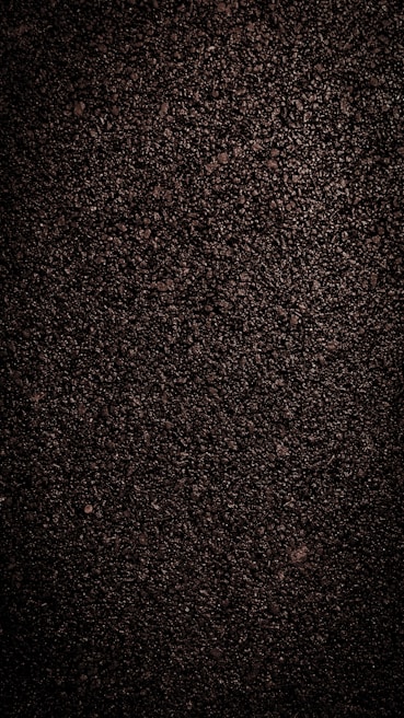 A close-up view of dark, coarse textured asphalt with a rough and uneven surface. The material appears dense with small stones and pebbles visible throughout.