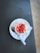 A glass jar filled with a dessert topped with fresh, sliced strawberries and what appears to be brown sugar or a crumb topping. It is placed on a white plate with a napkin beneath it, suggesting a clean presentation. A silver spoon is also on the plate, adding to the ready-to-eat appeal. The background is a smooth dark surface, contrasting with the vibrant colors of the dessert.