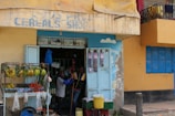 A small cereal shop with a weathered sign is visible. Outside, there is a fruit and vegetable stall displaying bananas, tomatoes, avocados, and other produce. Two people are interacting near the entrance, with a variety of goods visible through the open doors. The building exterior is painted in shades of yellow and blue, with a rough, urban aesthetic.