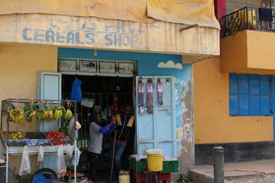 A small cereal shop with a weathered sign is visible. Outside, there is a fruit and vegetable stall displaying bananas, tomatoes, avocados, and other produce. Two people are interacting near the entrance, with a variety of goods visible through the open doors. The building exterior is painted in shades of yellow and blue, with a rough, urban aesthetic.