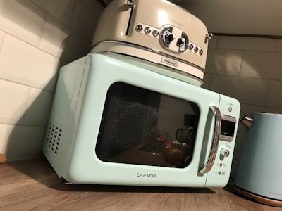 A retro-style kitchen setup featuring a pastel blue microwave and a cream-colored toaster stacked on top. Next to them is a matching pastel blue kettle. The appliances are placed on a wooden countertop against a white tiled wall.