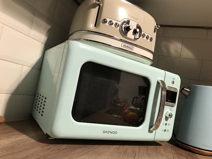 A retro-style kitchen setup featuring a pastel blue microwave and a cream-colored toaster stacked on top. Next to them is a matching pastel blue kettle. The appliances are placed on a wooden countertop against a white tiled wall.