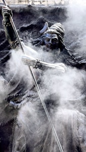 A detailed sculpture of a figure with an ornate headdress, holding a long staff or spear, enveloped in swirling mist or smoke. The intricate carvings and flowing robes add to the mystical and ancient appearance.