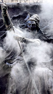 A detailed sculpture of a figure with an ornate headdress, holding a long staff or spear, enveloped in swirling mist or smoke. The intricate carvings and flowing robes add to the mystical and ancient appearance.