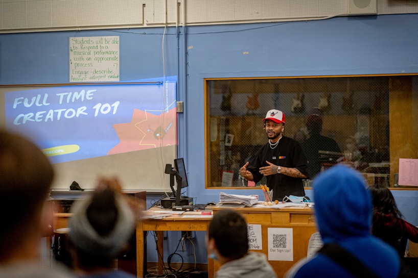 A person is standing at the front of a classroom, wearing a red and white cap and speaking to an audience. The screen behind them displays 'Full Time Creator 101.' The classroom setting includes a desk cluttered with papers, pens, and other objects. There is also a notice board and a window showing a reflection of the room.