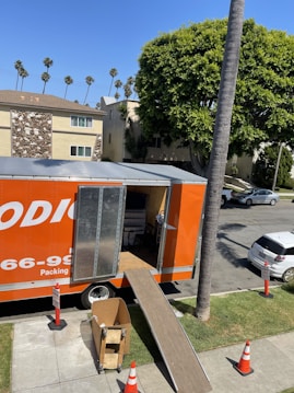 An orange moving truck is parked on a suburban street with its back door open. A ramp extends from the truck to the sidewalk, and there are traffic cones placed around the truck. Several cardboard boxes and items are visible inside the truck. Nearby, a tree provides shade, and apartment buildings line the street. There are also cars parked along the road.