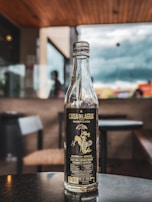 A clear glass bottle with a dark label stands on a reflective surface indoors, possibly a cafe or restaurant. The background is blurred, showing the outline of chairs, tables, and large windows with a cloudy sky visible outside.