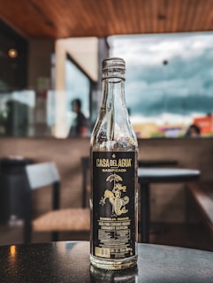 A clear glass bottle with a dark label stands on a reflective surface indoors, possibly a cafe or restaurant. The background is blurred, showing the outline of chairs, tables, and large windows with a cloudy sky visible outside.