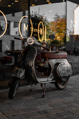 A classic, vintage scooter with a worn and rustic appearance is parked on a stone pavement. The scooter is positioned in front of a modern storefront with reflective glass, showcasing a blurred cityscape and trees reflected in the window. The signage on the window features bold white letters and circular designs, creating an urban and nostalgic feel.