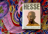 A book titled 'Tales of Student Life' by Hermann Hesse is placed on top of a vividly colored, intricately patterned fabric. The book cover features a stylized portrait of a man with glasses in muted colors, while the background fabric is adorned with bold, swirling patterns in pink, purple, blue, yellow, and red.