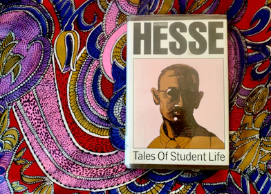 A book titled 'Tales of Student Life' by Hermann Hesse is placed on top of a vividly colored, intricately patterned fabric. The book cover features a stylized portrait of a man with glasses in muted colors, while the background fabric is adorned with bold, swirling patterns in pink, purple, blue, yellow, and red.
