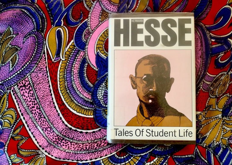 A book titled 'Tales of Student Life' by Hermann Hesse is placed on top of a vividly colored, intricately patterned fabric. The book cover features a stylized portrait of a man with glasses in muted colors, while the background fabric is adorned with bold, swirling patterns in pink, purple, blue, yellow, and red.
