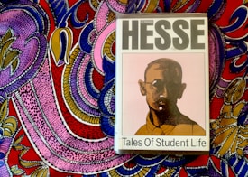 A book titled 'Tales of Student Life' by Hermann Hesse is placed on top of a vividly colored, intricately patterned fabric. The book cover features a stylized portrait of a man with glasses in muted colors, while the background fabric is adorned with bold, swirling patterns in pink, purple, blue, yellow, and red.