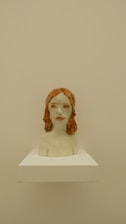 A ceramic bust sculpture of a person with red hair. The sculpture is placed on a white pedestal against a beige background. The facial features are somewhat abstract, and the surface appears to be glazed, giving it a shiny finish.