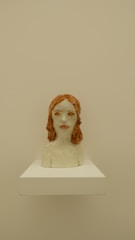 A ceramic bust sculpture of a person with red hair. The sculpture is placed on a white pedestal against a beige background. The facial features are somewhat abstract, and the surface appears to be glazed, giving it a shiny finish.