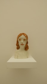 A ceramic bust sculpture of a person with red hair. The sculpture is placed on a white pedestal against a beige background. The facial features are somewhat abstract, and the surface appears to be glazed, giving it a shiny finish.