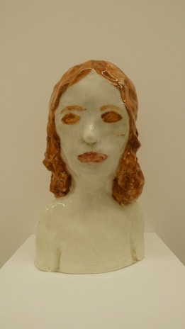 A ceramic bust of a person with long, reddish-brown hair and a neutral expression. The piece is simplistic in detail, focusing on the basic shapes and textures to convey the human form. The ceramic has a glossy finish, highlighting the smoothness and the subtle curves of the sculpture.