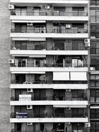 Tall residential building showcasing multiple balconies across several floors. Each balcony is outfitted with air conditioning units, railings, and an array of plants and outdoor furniture. Signs with contact information suggest apartments available for sale. The structure is composed of brick and concrete, exhibiting a functional architectural design.