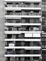Tall residential building showcasing multiple balconies across several floors. Each balcony is outfitted with air conditioning units, railings, and an array of plants and outdoor furniture. Signs with contact information suggest apartments available for sale. The structure is composed of brick and concrete, exhibiting a functional architectural design.
