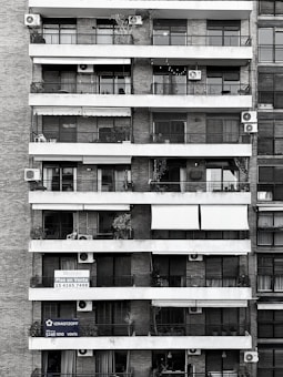 Tall residential building showcasing multiple balconies across several floors. Each balcony is outfitted with air conditioning units, railings, and an array of plants and outdoor furniture. Signs with contact information suggest apartments available for sale. The structure is composed of brick and concrete, exhibiting a functional architectural design.