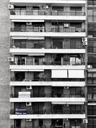 Tall residential building showcasing multiple balconies across several floors. Each balcony is outfitted with air conditioning units, railings, and an array of plants and outdoor furniture. Signs with contact information suggest apartments available for sale. The structure is composed of brick and concrete, exhibiting a functional architectural design.