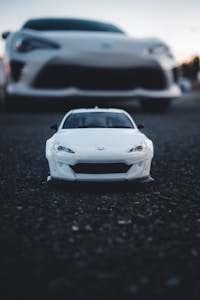 A small scale model of a white sports car is positioned in the foreground on a dark, textured surface, with a full-sized car of the same model blurred in the background.