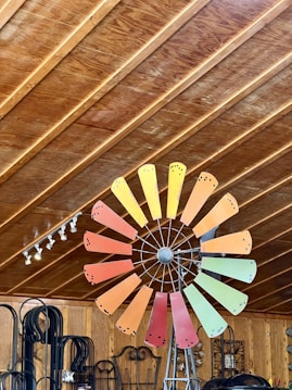 A colorful metal windmill with blades in various shades including yellow, orange, red, and green is situated indoors against a wooden paneled ceiling. The setting appears to be a display or store environment with other metal items visible in the background.