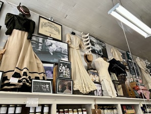 An assortment of vintage clothing and framed photographs displayed on a high shelf. The clothing includes long dresses in cream and black with lace and frilled details, alongside various hats and an umbrella. Numerous black-and-white photographs are interspersed, featuring groups and individuals in historical attire. Shelving beneath holds jars of preserves and other items, suggesting a museum or antique store setting.
