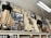 An assortment of vintage clothing and framed photographs displayed on a high shelf. The clothing includes long dresses in cream and black with lace and frilled details, alongside various hats and an umbrella. Numerous black-and-white photographs are interspersed, featuring groups and individuals in historical attire. Shelving beneath holds jars of preserves and other items, suggesting a museum or antique store setting.