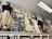 An assortment of vintage clothing and framed photographs displayed on a high shelf. The clothing includes long dresses in cream and black with lace and frilled details, alongside various hats and an umbrella. Numerous black-and-white photographs are interspersed, featuring groups and individuals in historical attire. Shelving beneath holds jars of preserves and other items, suggesting a museum or antique store setting.