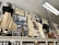 An assortment of vintage clothing and framed photographs displayed on a high shelf. The clothing includes long dresses in cream and black with lace and frilled details, alongside various hats and an umbrella. Numerous black-and-white photographs are interspersed, featuring groups and individuals in historical attire. Shelving beneath holds jars of preserves and other items, suggesting a museum or antique store setting.