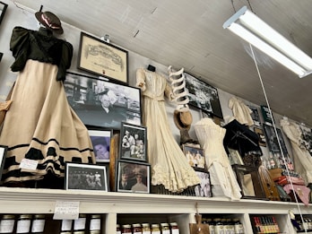 An assortment of vintage clothing and framed photographs displayed on a high shelf. The clothing includes long dresses in cream and black with lace and frilled details, alongside various hats and an umbrella. Numerous black-and-white photographs are interspersed, featuring groups and individuals in historical attire. Shelving beneath holds jars of preserves and other items, suggesting a museum or antique store setting.