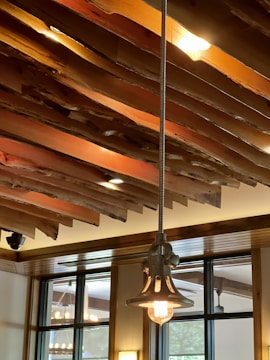 A modern interior space featuring a ceiling with artistic, layered wooden beams and a hanging industrial-style light fixture. Natural light illuminates the room through large windows, highlighting the wooden textures and metal elements.